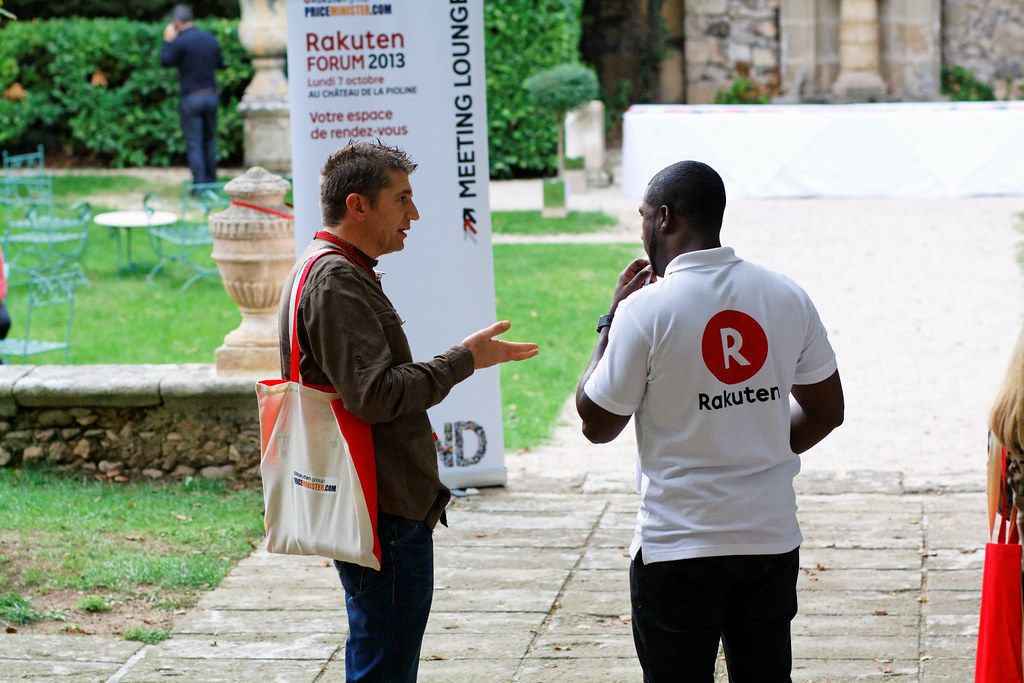 라쿠텐 포럼 2013 행사장에서 라쿠텐 로고 티셔츠를 입은 직원과 참가자가 영어로 대화하는 모습